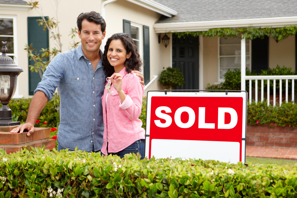 A Hispanic couple posing in the front yard with a sold sign